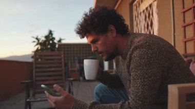 Gülümseyen genç adam terasta oturuyor, cep telefonu kullanıyor.