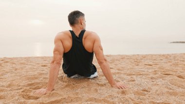 Orta yaşlı atletik adam antrenmandan sonra sahilde oturur ve denize bakar.