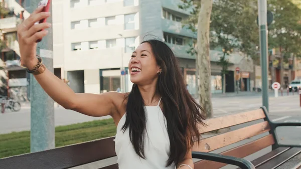Beyaz bluzlu güzel esmer kız gülümsüyor ve kentin arka planında bankta otururken selfie çekiyor.