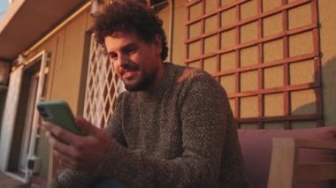 Gülümseyen genç adam terasta oturuyor, cep telefonu kullanıyor.