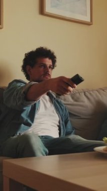 Genç adam elinde uzaktan kumandayla oturma odasında oturuyor.