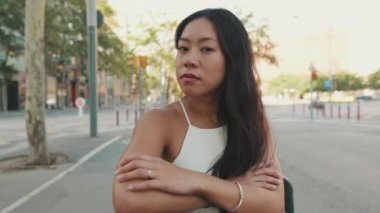 Genç bir kadın kollarını kavuşturup bankta oturur yan tarafa bakar, başını çevirir ve kameraya bakar.