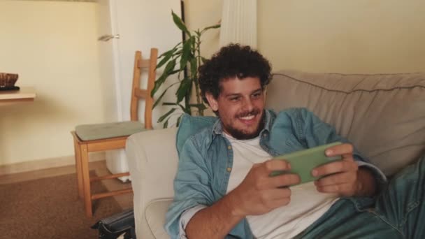 Sorrindo Jogador Cara Joga Jogos Vídeo Com Telefone Móvel Deitado — Vídeo de Stock