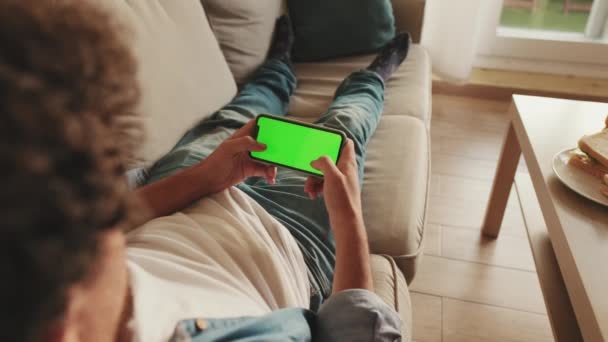 若いです男横たわっソファ再生上の携帯電話で緑のモックアップ画面クロマキー — ストック動画