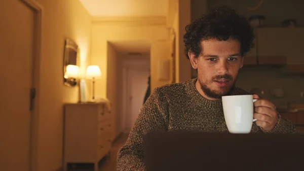 stock image Guy works on laptop and drinks coffee sitting at table in the kitchen in the evening, freelancer working from home, working on the Internet, using social networks