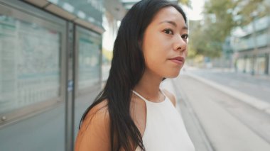 Beyaz bluz ve kot giyen esmer kızın toplu taşıma durağında dikilip etrafa bakındığı yakın plan.