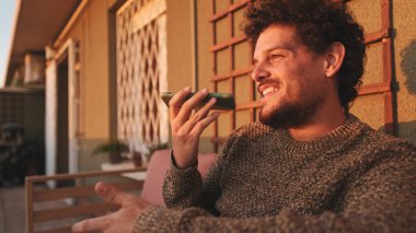 Terasta oturan ve hoparlörle akıllı telefondan konuşan mutlu adam.