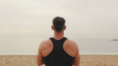 Atletik olgun bir adam sabah plajda dikilip rahatlama egzersizleri yapıyor.