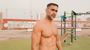 Atletik olgun bir adam sabah spor sahasında dikilirken kameraya bakıyor.