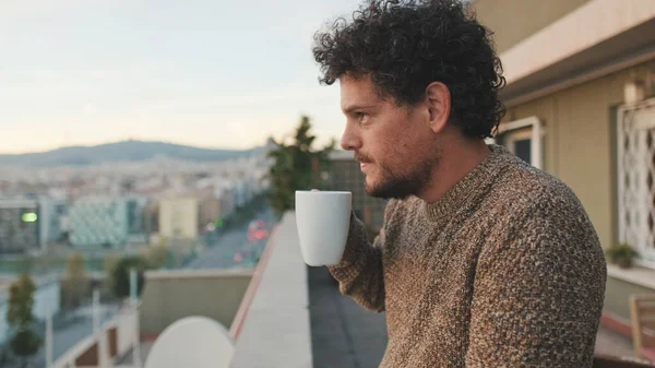 stock image Peaceful guy looks at the city from the balcony and drinks coffee, enjoys the beautiful view