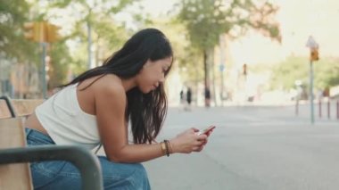 Genç kadın bankta oturuyor ve cep telefonu kullanıyor.
