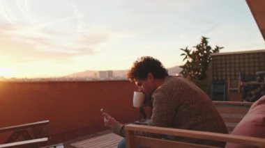 Gülümseyen genç adam terasta oturuyor, cep telefonu kullanıyor.