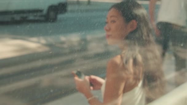 若い女性は公共交通機関の停留所に座って 携帯電話を使用して ガラスを通してぼやけた景色 — ストック動画