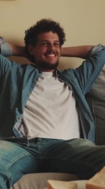 Oturma odasında oturup televizyon izleyen, öğle yemeği yiyen genç adam.