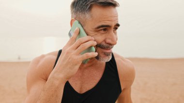 Yaklaş, orta yaşlı spor kıyafetli kaslı adam deniz kenarında gezinirken akıllı telefondan konuşuyor.