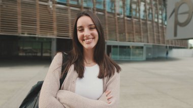 Girl student stands outside the university building crosses her arms, looks at the camera and smiles