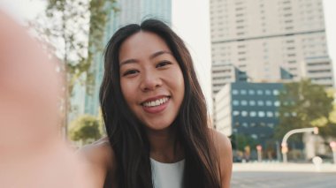 Yaklaş, beyaz bluz giyen sevimli mutlu kız ve modern şehir arka planında dururken kotlar cep telefonuyla selfie çekiyor.