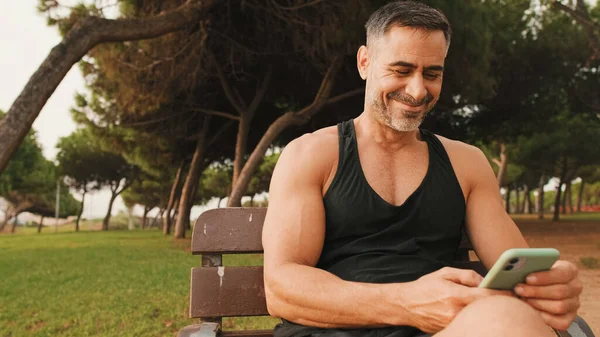 Smiling middle aged muscular man in sportswear typing messages on mobile phone while sitting in park after workout