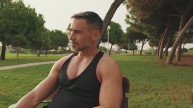 Spor giyimli orta yaşlı bir adam parkta otururken dinlenmeyi sever.