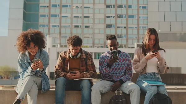 Happy group of friends using phone and smiling together outside. Students sit next to the university building looking at their mobile phones and talking