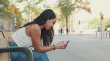 Genç kadın bankta oturuyor ve cep telefonu kullanıyor.