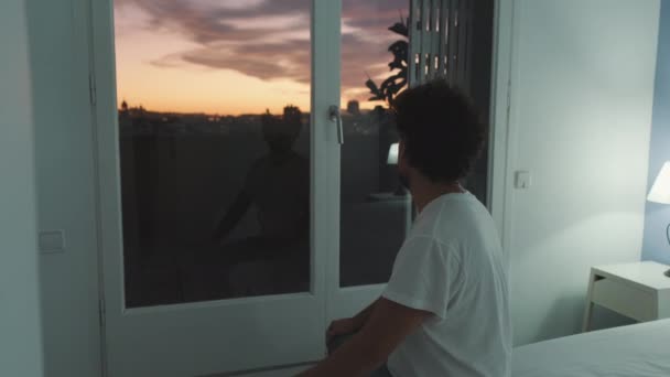 若いです男に座ってベッドルームの窓の外を見て夕日 — ストック動画