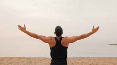 Atletik olgun bir adam sabah plajda dikilip rahatlama egzersizleri yapıyor.