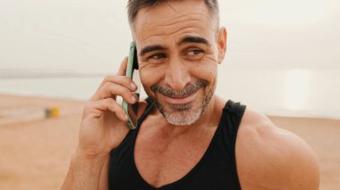 Yaklaş, orta yaşlı spor kıyafetli kaslı adam deniz kenarında gezinirken akıllı telefondan konuşuyor.