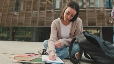 Öğrenci dışarıda otururken ders çalışıyor. Kız sınava hazırlanıyor, not defterine not alıyor.