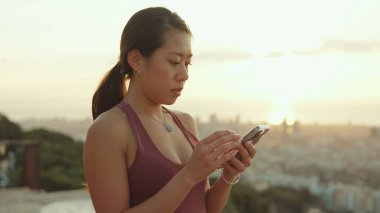 Clopse up, happy athletic woman using mobile phone while standing at lookout point at dawn