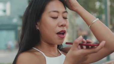 Yakından, bankta oturan genç kadın cep telefonuyla sesli mesaj gönderiyor.