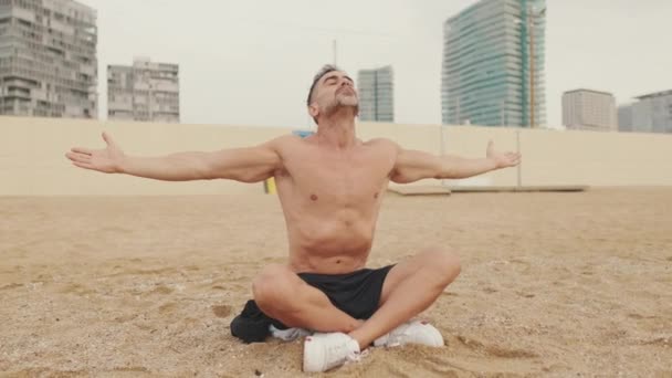 成熟した男は 海岸で砂の上に座って呼吸演習を行う — ストック動画