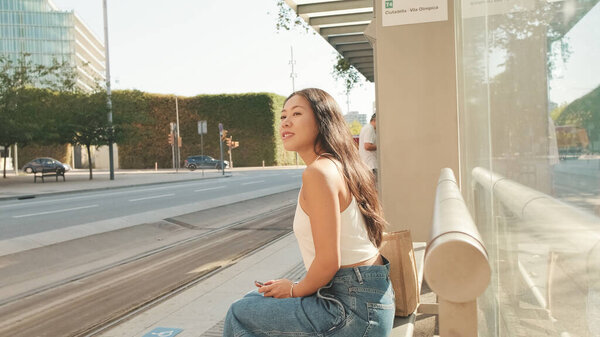 Pretty brunette girl, dressed in white top and jeans, sits at public transport stop with smartphone in her hands, looks around