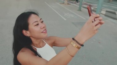 Genç bir kadın modern şehir arka planında dururken cep telefonuyla selfie çekiyor.