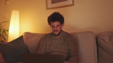 Genç adam oturma odasında kanepede dizüstü bilgisayarla oturuyor.