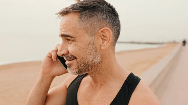 stock image Close up, middle-aged muscular man in sportswear talking from smartphone while walking on promenade on sea background