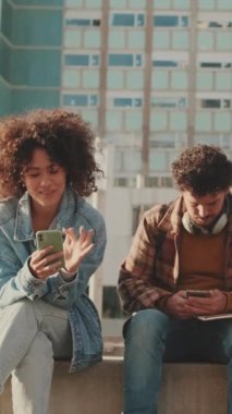 Couple of students are using mobile phones and talking while sitting outside