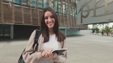 Elinde defterleri olan bir kız öğrenci üniversite binasının önünde durmuş kameraya bakıyor ve gülümsüyor.