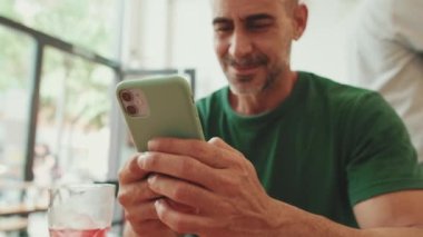 Orta yaşlı bir adam kafede oturup cep telefonundan video izliyor.