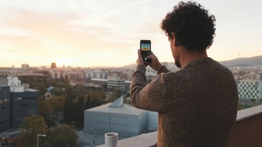 Mutlu adam şafakta balkonda duruyor, fotoğraf çekiyor, akıllı telefondaki güzel manzaranın videosunu çekiyor.