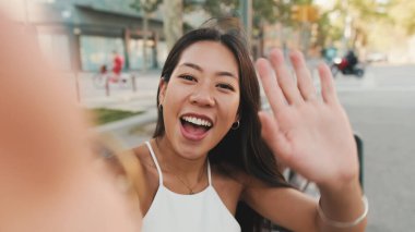 Beyaz bluzlu güzel esmer kız gülümsüyor ve kentin arka planında bankta otururken selfie çekiyor.