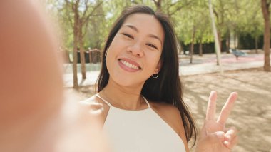 Beyaz bluz ve kot giyen gülümseyen esmer kız, şehir parkında dururken cep telefonundan selfie çekiyor.