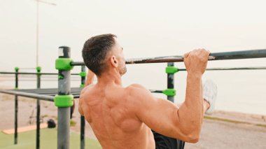Orta yaşlı kaslı bir adam deniz arka planındaki yatay çubukta egzersiz yapıyor.
