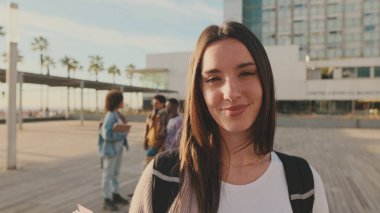 Kız kameraya bakarken gülümsüyor. Elinde not defterleri olan bir öğrenci kameraya bakar ve gülümser.