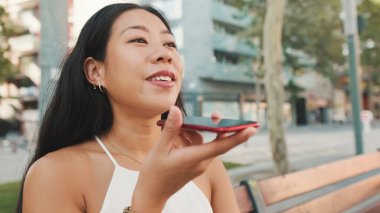 Yaklaş, beyaz bluz ve kot giyen tatlı esmer kız, kentin arka planında bankta oturuyor, akıllı telefondan sesli mesaj gönderiyor.