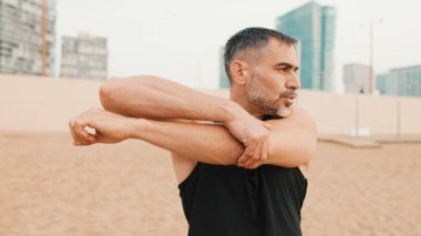 Atletik olgun bir adam sabahları kumsalda dururken kol kaslarını esnetir.