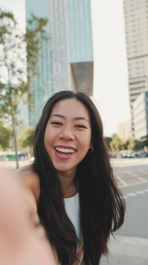 Yaklaş, beyaz bluz giyen sevimli mutlu kız ve modern şehir arka planında dururken kotlar cep telefonuyla selfie çekiyor.