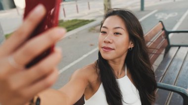 Beyaz bluzlu güzel esmer kız gülümsüyor ve kentin arka planında bankta otururken selfie çekiyor.