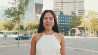 Yakın plan güzel, mutlu, beyaz bluzlu ve kot pantolonlu esmer kız modern şehir arka planına bakıyor.