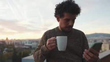 Genç adam balkonda duruyor, cep telefonu kullanıyor.